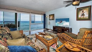 A spacious living room with a large window overlooking the ocean, wicker furniture, and a ceiling fan.
