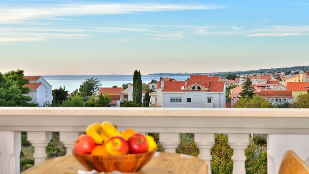 A balcony with a wooden table holding a fruit bowl, overlooking a coastal town with red-roofed houses and a view of the sea. - Ina Apartments (Zadar)