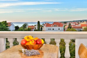A balcony with a wooden table holding a fruit bowl, overlooking a coastal town with red-roofed houses and a view of the sea. - Ina Apartments (Zadar)