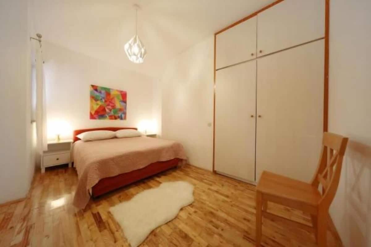 A bedroom with a bed, a wardrobe, a chair, a rug, and a painting on the wall.