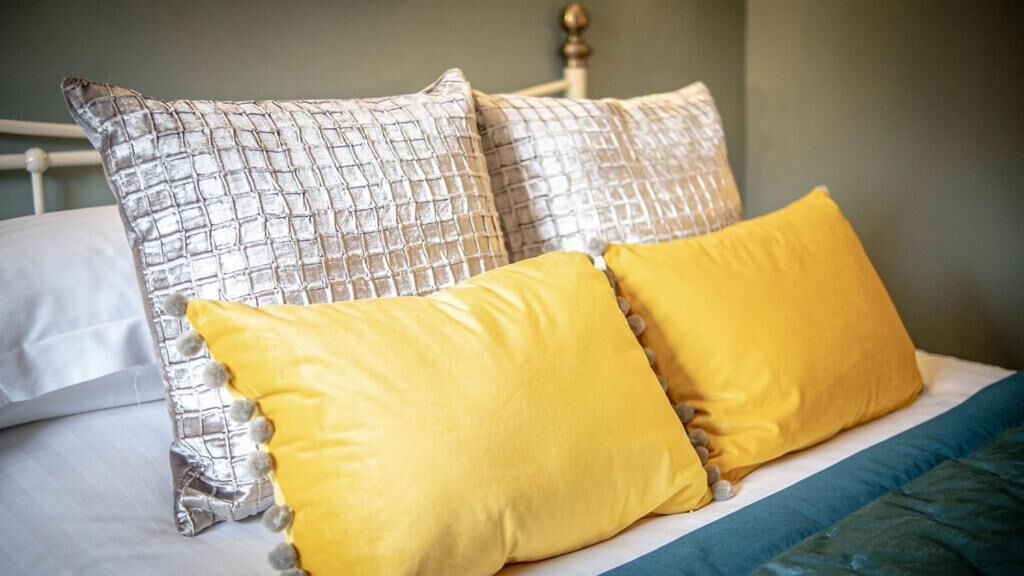 A bed with two pillows, one silver and one yellow, and a blue blanket.