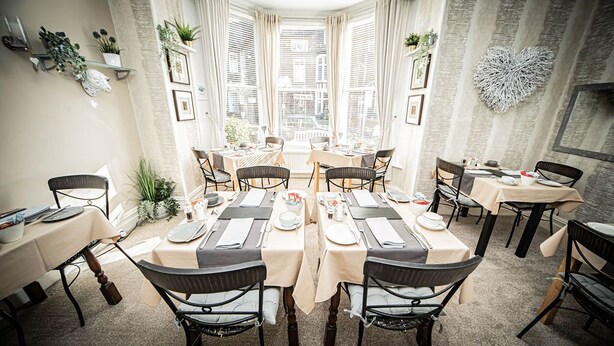 A dining area with multiple tables set for meals, large windows with curtains, and decorative wall elements. - Hillsdale in Ambleside (Ambleside)
