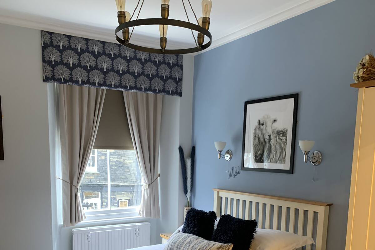 A bedroom with a bed, a nightstand, a chair, a window with curtains, a chandelier, wall art, and a radiator.