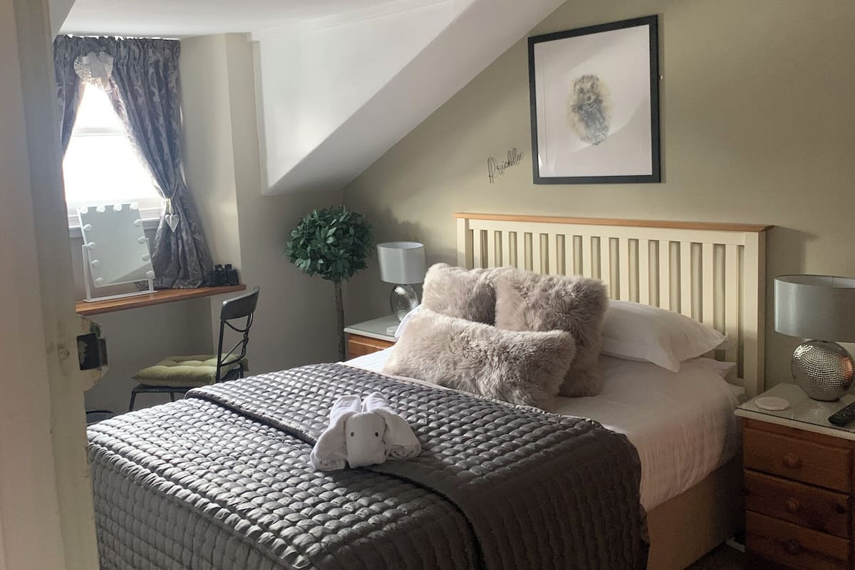 A bedroom with a sloped ceiling, a bed with a fluffy pillow, a bedside table with a lamp, and a framed picture on the wall.