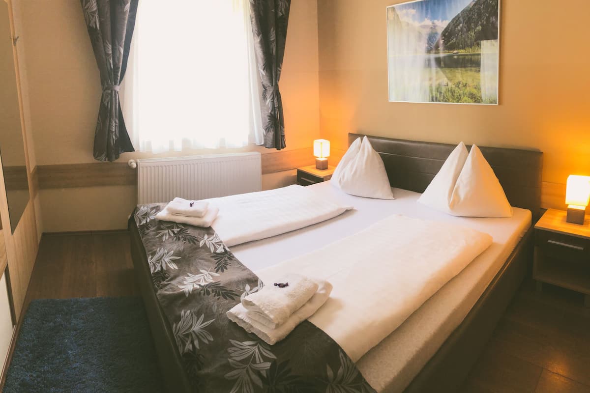 Habitación de hotel con una cama, dos lámparas de noche, un ventanal con cortinas, un radiador y un cuadro en la pared.
