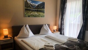 A hotel room with a bed, two bedside tables with lamps, a painting of a mountain landscape, and a window with curtains. - Hotel Mallnitz Appartements (Mallnitz)