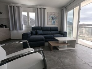 Un salon moderne avec un canapé bleu foncé, une table basse blanche et une vue sur un paysage montagneux grâce à de grandes fenêtres.