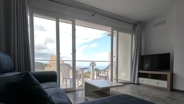Un soggiorno moderno con un divano, una TV su un mobile in legno e una porta scorrevole in vetro che dà su un balcone con mobili da esterno.