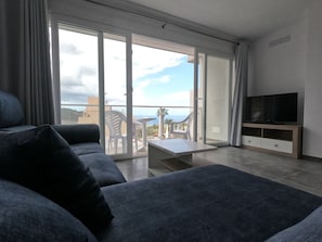 Un salon moderne avec un canapé, un téléviseur posé sur un meuble en bois et une porte-fenêtre coulissante donnant sur un balcon aménagé avec du mobilier d’extérieur.