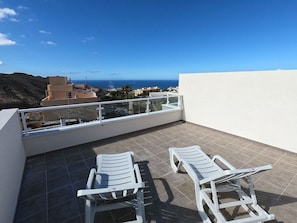 A balcony with two white lounge chairs, a tiled floor, and a view of the sea and buildings in the distance.