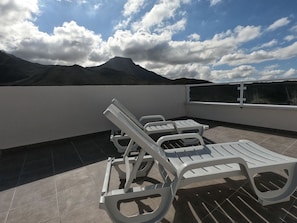 A rooftop terrace with white lounge chairs, a view of mountains, and a clear sky.