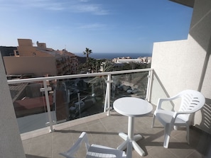 Un balcón con barandilla de vidrio, una pequeña mesa redonda, dos sillas de plástico blanco y vistas al mar y a edificios.