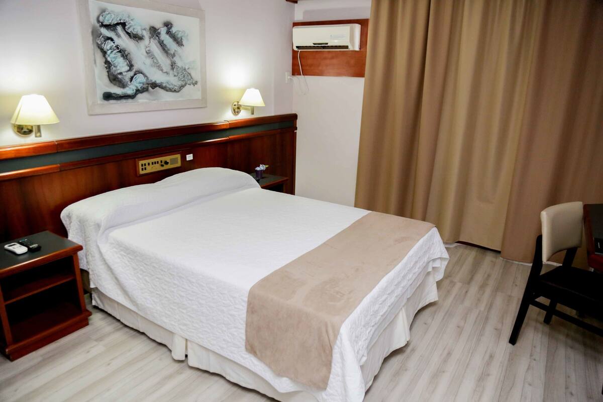 Une chambre d’hôtel comprenant un lit, des lampes de chevet, une table de chevet, une chaise, un climatiseur fixé au mur et une fenêtre avec des rideaux.