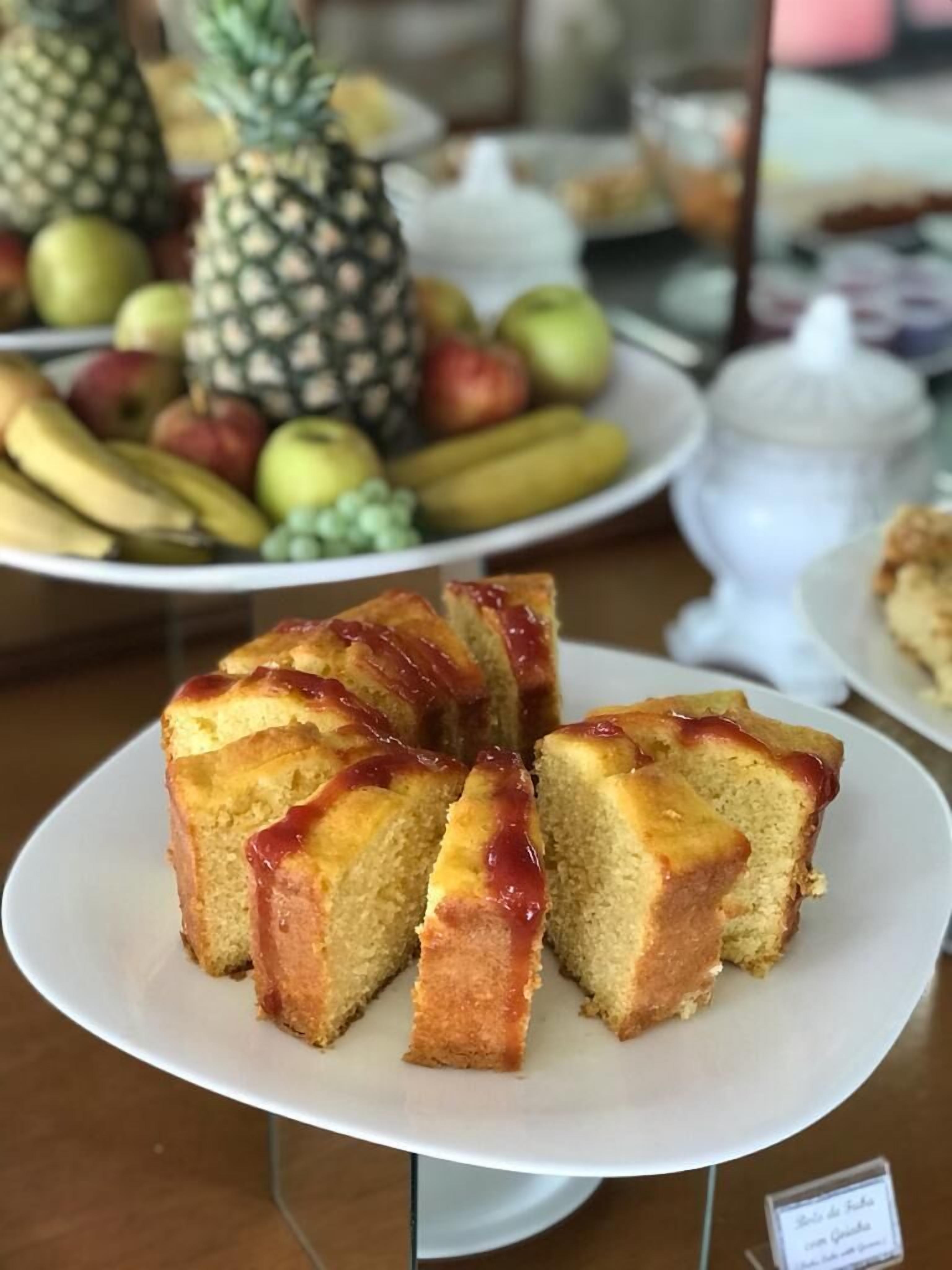 Bolo com cobertura sobre um prato branco, com uma bandeja de frutas ao fundo.