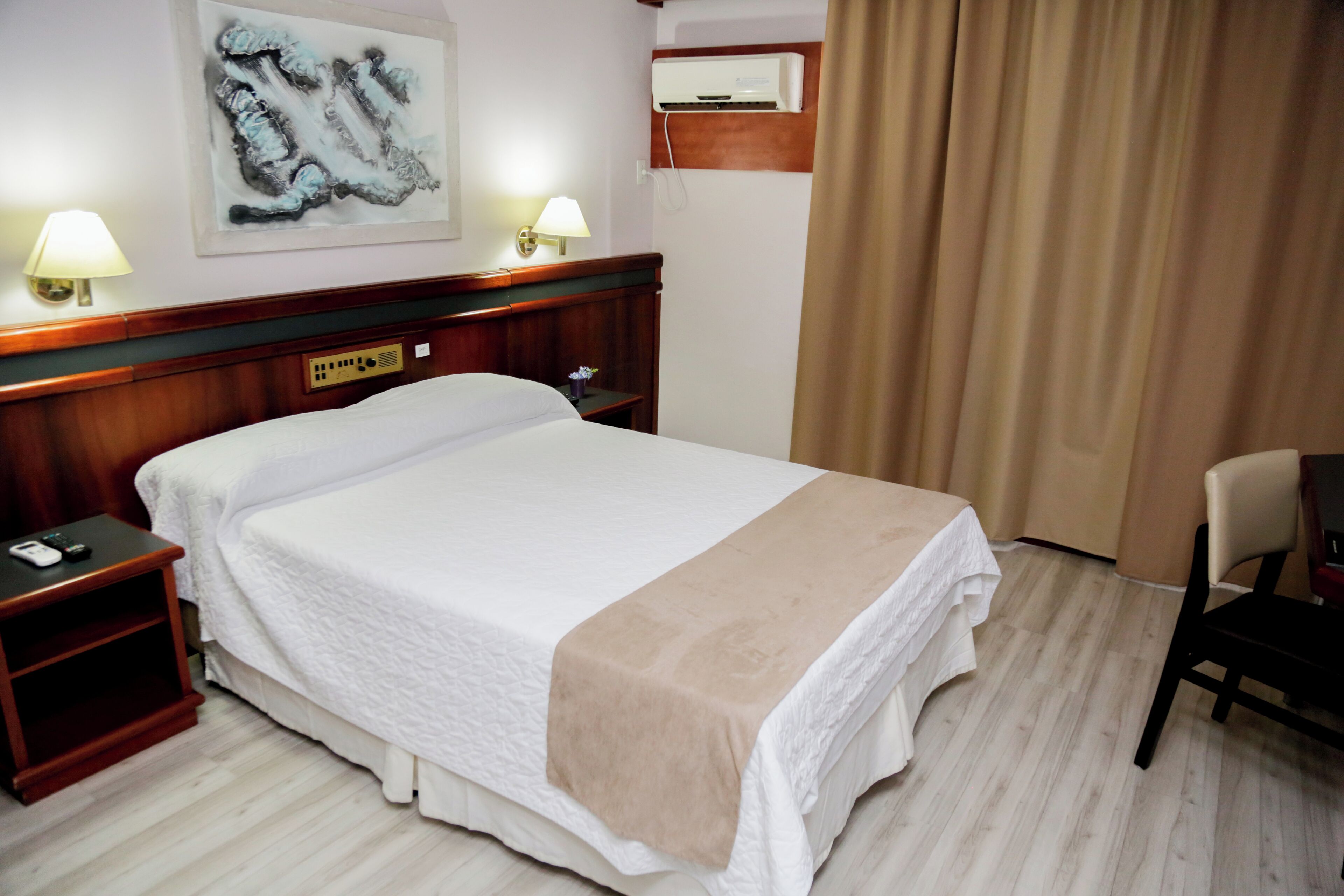 A hotel room with a large bed, bedside lamps, a wooden headboard, a wall-mounted air conditioner, a small table with a lamp, and a chair.