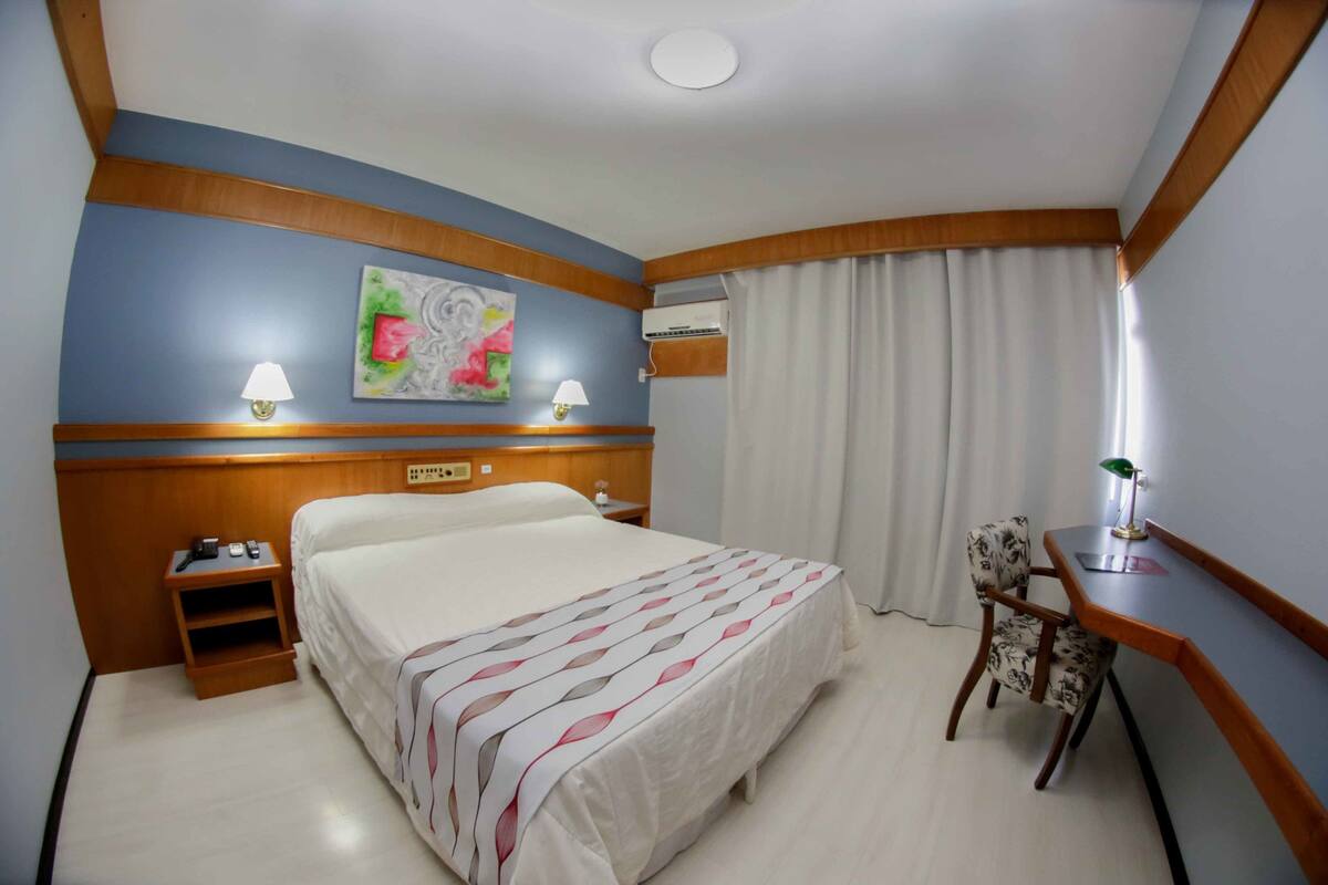 Une chambre d’hôtel comprenant un lit, des tables de chevet, un bureau avec une chaise, un climatiseur fixé au mur et un tableau.