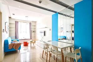 Uma sala de estar moderna com um sofá azul, uma mesa de jantar de vidro e cadeiras brancas. Há uma janela grande com cortinas, uma TV montada na parede e uma estante.