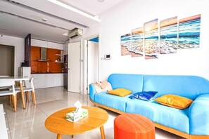 Uma sala de estar moderna com um sofá azul, uma mesa de centro de madeira e uma área de cozinha ao fundo.