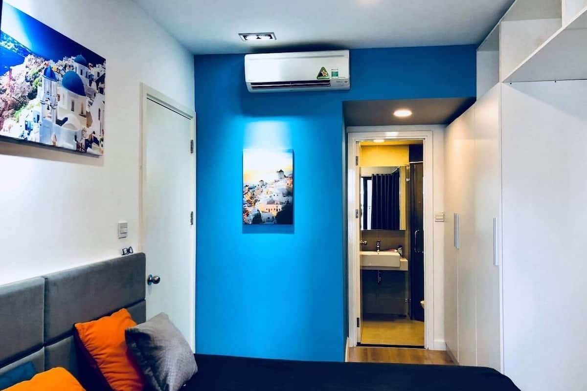 A compact hotel room with a bed, a blue accent wall, an air conditioning unit, a door leading to a bathroom, and a framed picture on the wall.