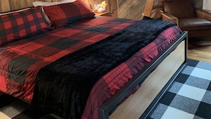 Un dormitorio con techo de madera inclinado, una cama con colcha de cuadros, una mesita de noche con lámpara y una escalera de madera.