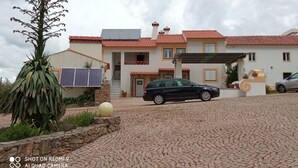 Une maison à deux étages avec un toit recouvert de tuiles rouges, une voiture garée dans l’allée et une grande plante au premier plan.