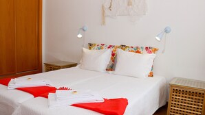 Un lit bien fait, avec du linge de lit blanc, un plaid rouge et une serviette pliée posée sur le côté. Il y a deux tables de chevet, l’une avec une lampe et l’autre avec un petit objet décoratif.