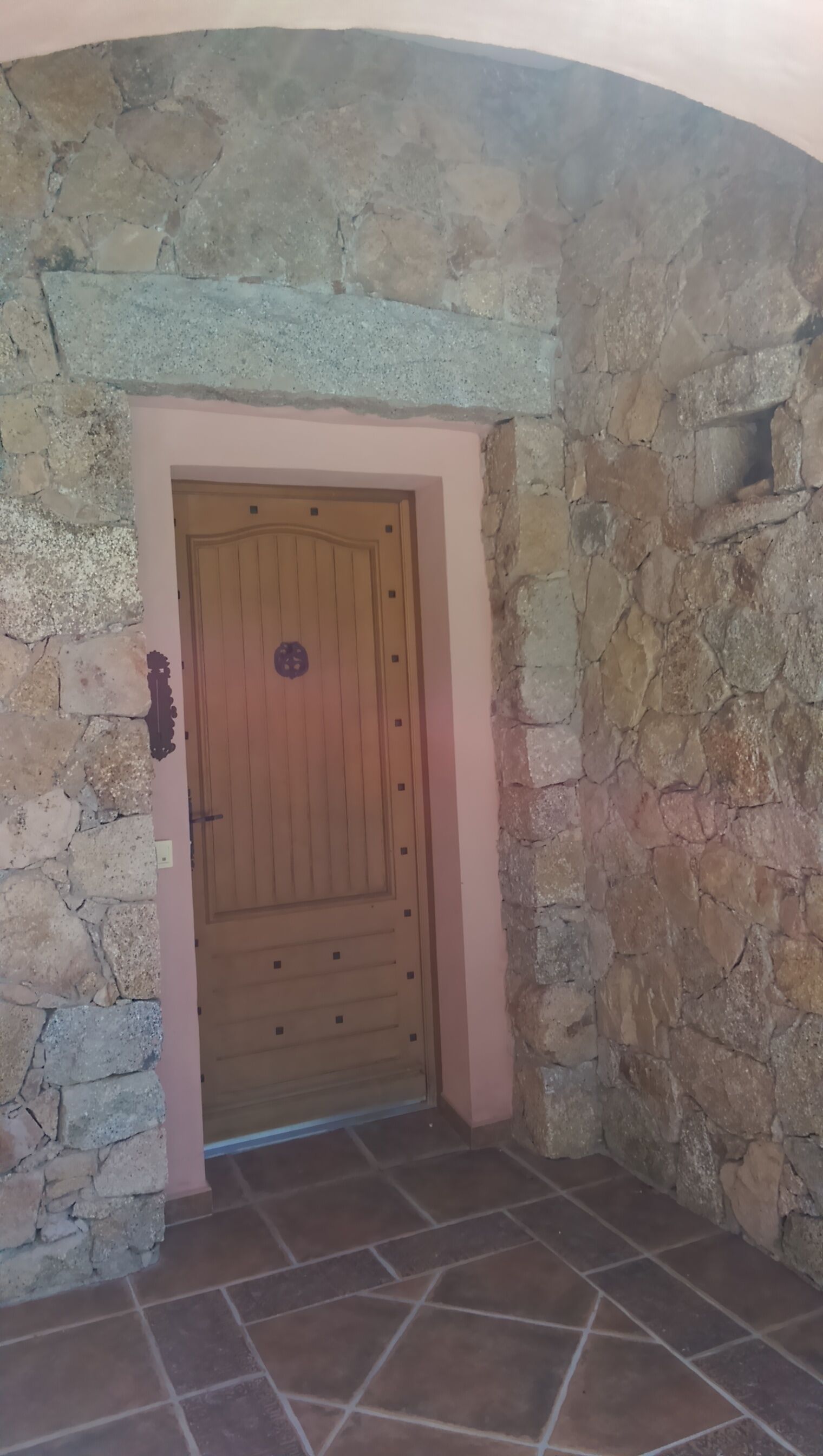 A stone-walled entrance with a wooden door, a small shelf, and tiled flooring.