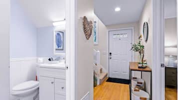Een badkamer met toilet, wastafel en spiegel. Er is een deur naar een andere kamer met een houten vloer en een klein tafeltje met een plant.