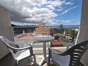 A balcony with two white plastic chairs, a small round table, and a view of a coastal road with buildings and palm trees. - Holiday apartment Adeje 14 (Adeje)