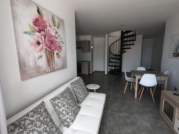Un salon moderne avec un canapé blanc, un coin repas avec une table et des chaises en bois, et un escalier menant à un niveau supérieur.
