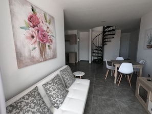 A modern living room with a white sofa, a dining area with a wooden table and chairs, and a staircase leading to an upper level.