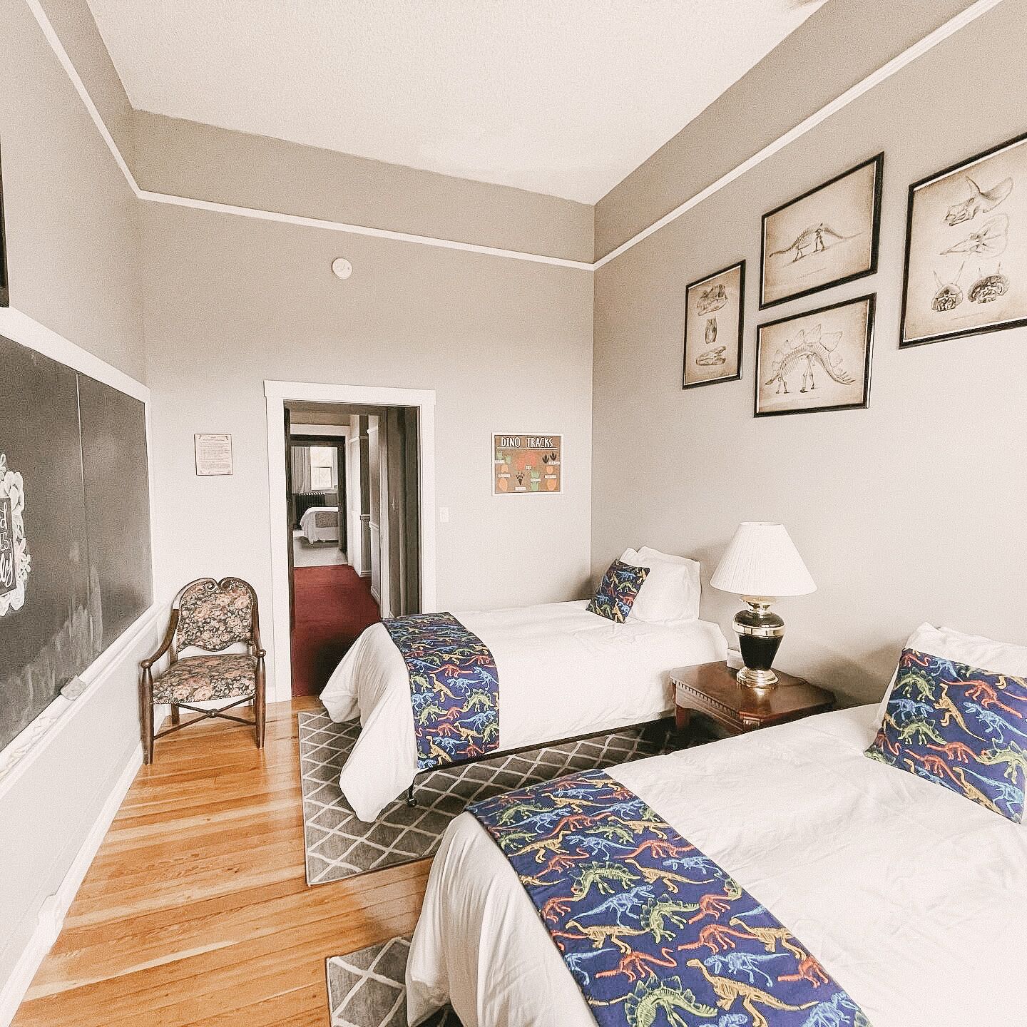 A hotel room with two beds, a chair, a desk with a lamp, and framed pictures on the wall.