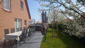 Terassi, jossa on pitkä pöytä ja tuoleja, katettu oleskelualue sekä puutarha, jossa on kukkivia puita.