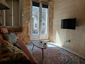 Ein Wohnzimmer mit einer Couch, einem kleinen Tisch, einem an der Wand befestigten Fernseher und einem Fenster mit Blick auf Gebäude.