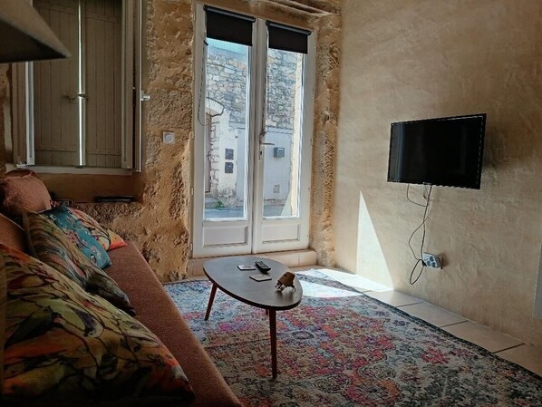 Ein Wohnzimmer mit einer Couch, einem kleinen Tisch, einem an der Wand befestigten Fernseher und einem Fenster mit Blick auf Gebäude.