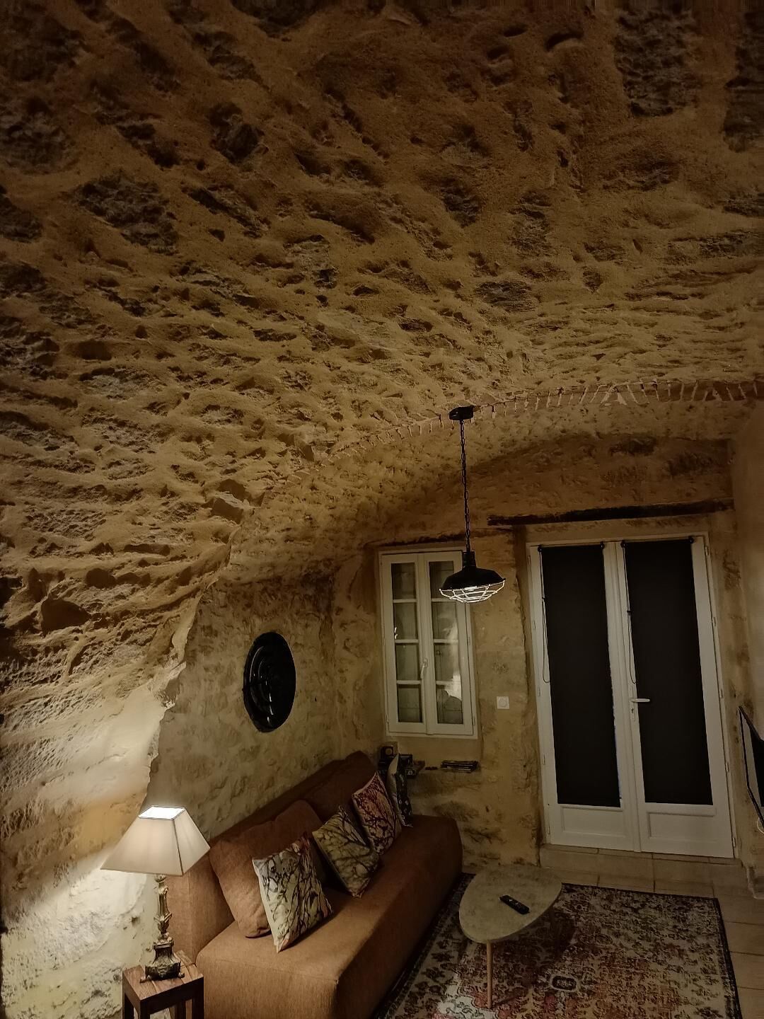 Een rustieke kamer met een stenen plafond, een bank met kussens, een tafel met een lamp en een raam met een deur.