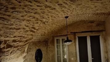 Een rustieke kamer met een stenen plafond, een bank met kussens, een tafel met een lamp en een raam met een deur.