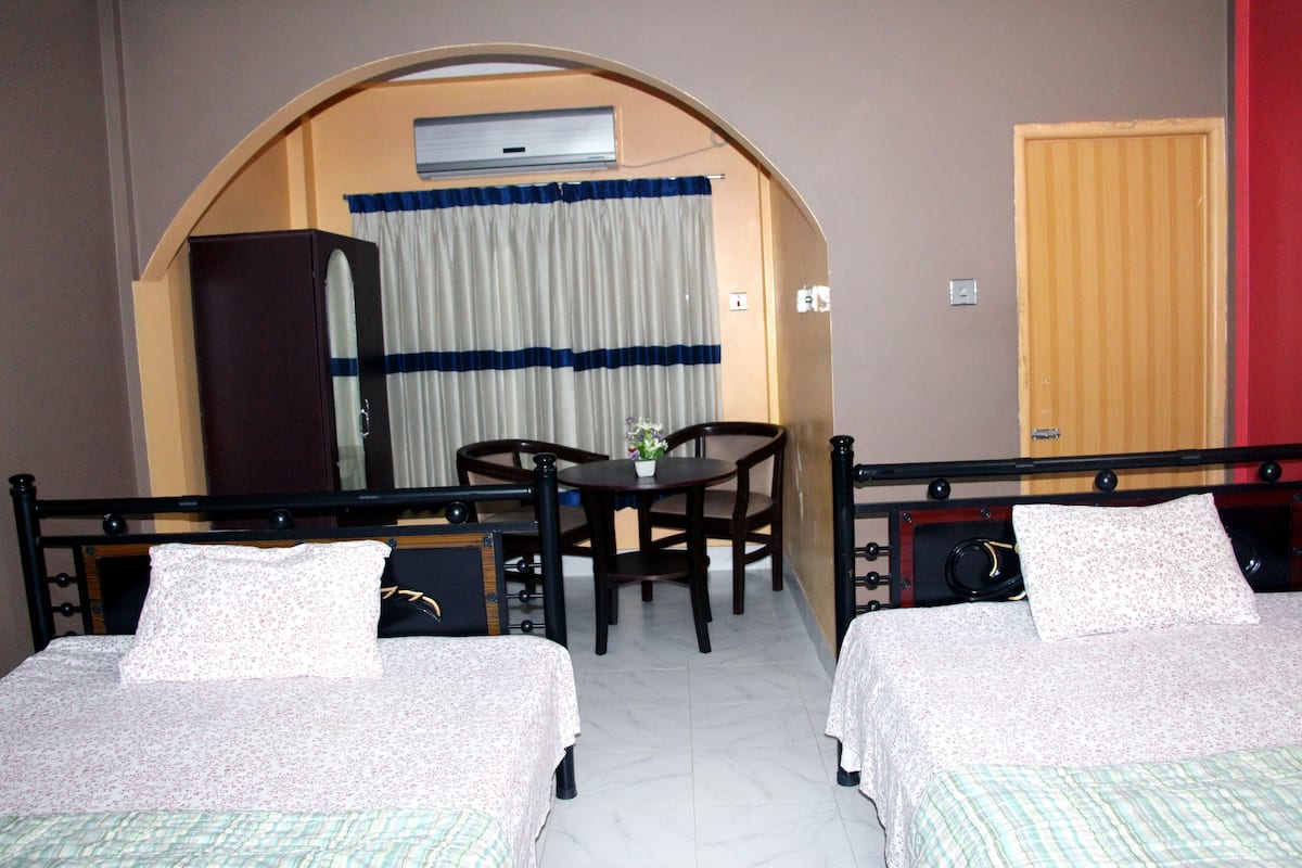 A hotel room with two beds, a dining table, chairs, a small table with a flower vase, and an air conditioning unit.
