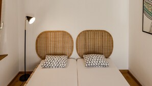 A room with a large woven ceiling light fixture, two wicker headboards with patterned cushions, a floor lamp, and a white bedspread.