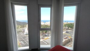 Ein Zimmer mit drei großen Fenstern, einem roten Kissen und Blick auf Stadt und Küste.