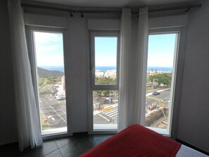 A room with three large windows, a red cushion, and a view of a cityscape and coastline.