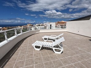 A rooftop terrace with white sun loungers, a tiled floor, and a view of the cityscape.