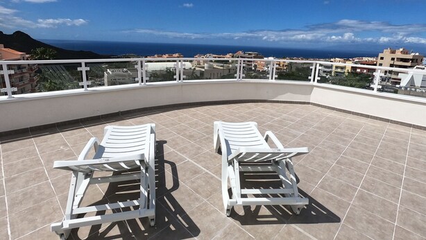 Eine Dachterrasse mit zwei weißen Liegestühlen, gefliestem Boden, einem weißen Geländer und Blick auf das Meer und Gebäude.