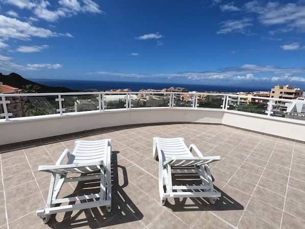 A rooftop terrace with two white lounge chairs, a tiled floor, a white railing, and a view of the sea and buildings.