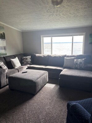 A living room with a large window, a sofa, an ottoman, and a remote control on the ottoman. - Lake Winnebago Vacation Rentals (Neenah)