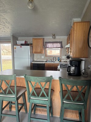 A kitchen with wooden cabinets, a refrigerator, a coffee maker, and a dining area with wooden chairs. - Lake Winnebago Vacation Rentals (Neenah)