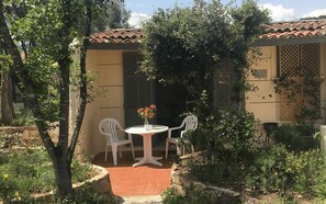 En hyggelig terrasse med et rundt bord og to stole, omgivet af grøn bevoksning og en bygning med et tag af fliser.