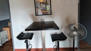 Un petit coin repas avec une table sombre, deux tabourets de bar noirs, un verre et un ventilateur sur pied.