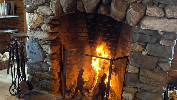 Une cheminée en pierre avec un feu qui crépite à l’intérieur, un chien allongé sur le sol devant, et une étagère en bois au-dessus de la cheminée.