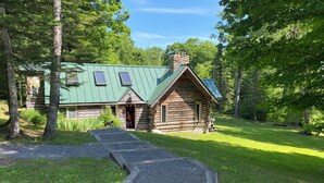 Une cabane rustique en rondins de bois, avec un toit végétalisé, une cheminée en pierre et un porche en bois.